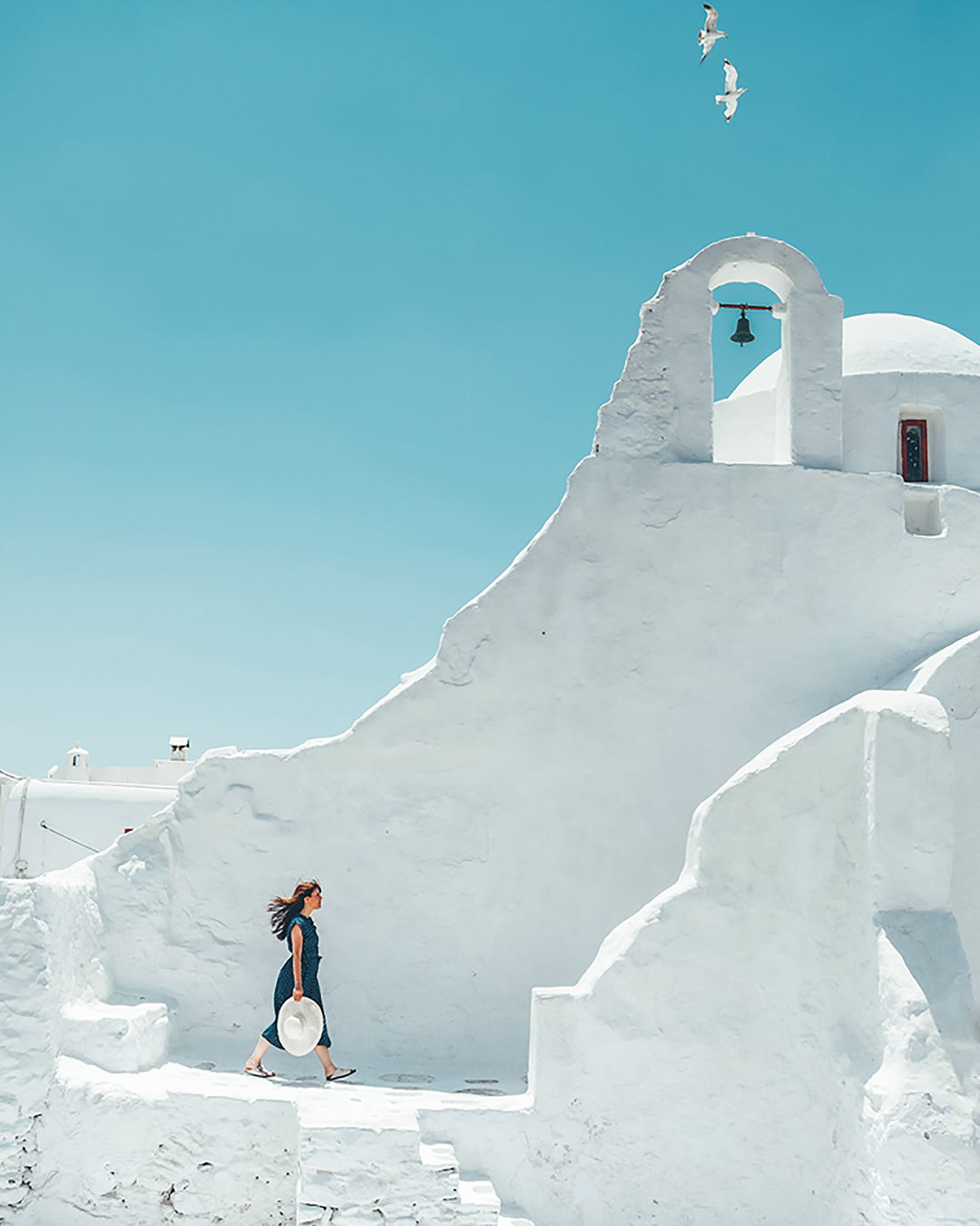 photo of a girl walking at panagia paraportiani with blew dress holding a hat